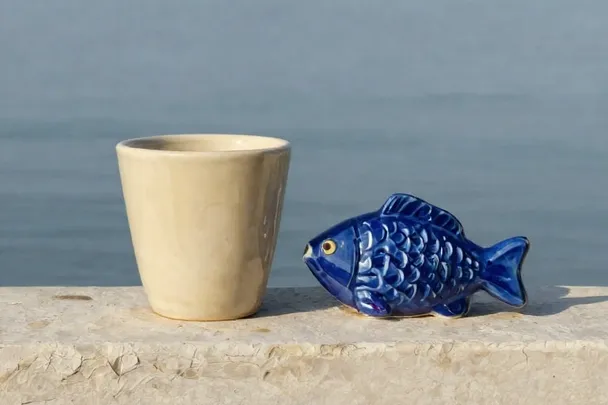 A ceramic cup stands beside a small fish-shaped sculpture near calm water.