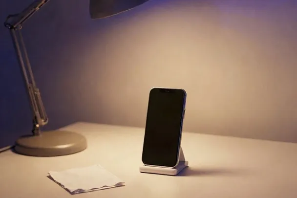 A phone on a small stand faces a clear cream surface beneath a soft desk lamp.